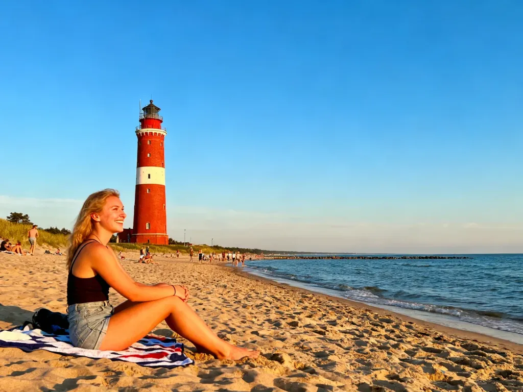 Uśmiechnięta kobieta wypoczywa na słonecznej plaży w Niechorzu, w tle widoczna charakterystyczna latarnia morska.